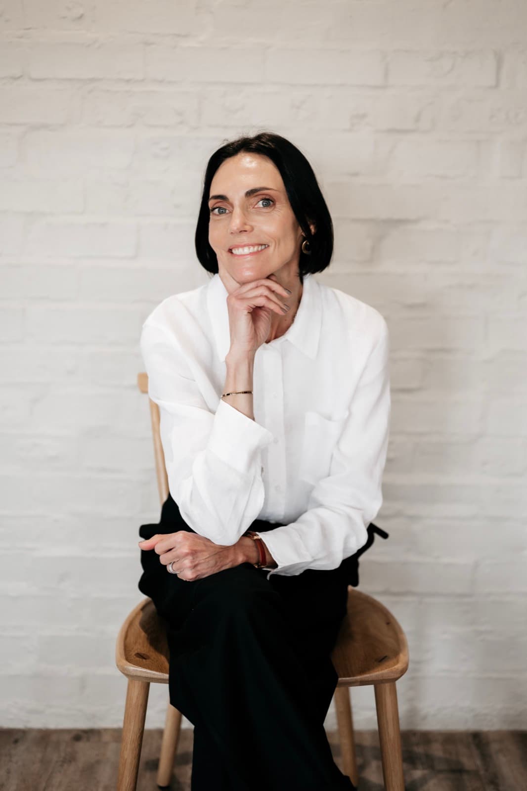 Belinda seated on a wooden stool in a crisp white shirt, hand resting at her chin